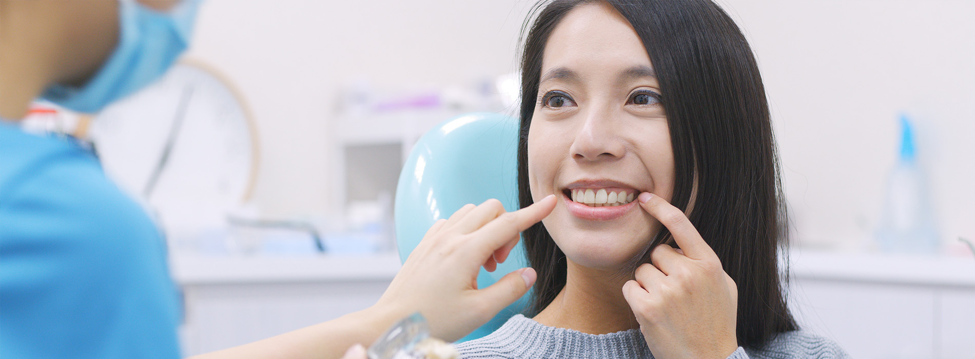 The image shows a woman sitting in a dental chair with a dentist performing an oral procedure on her, while she smiles at the camera.