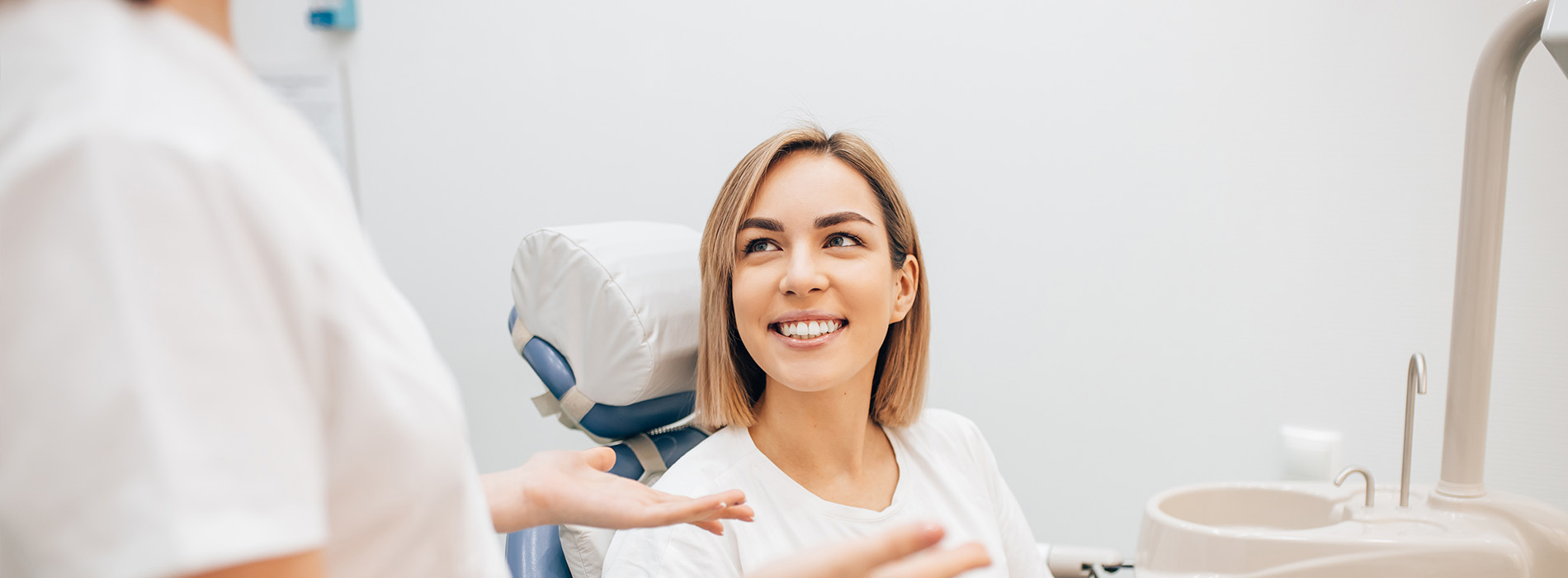 The image depicts a woman sitting in a dental chair with a smile on her face, being attended to by a dental professional who appears to be a dentist.