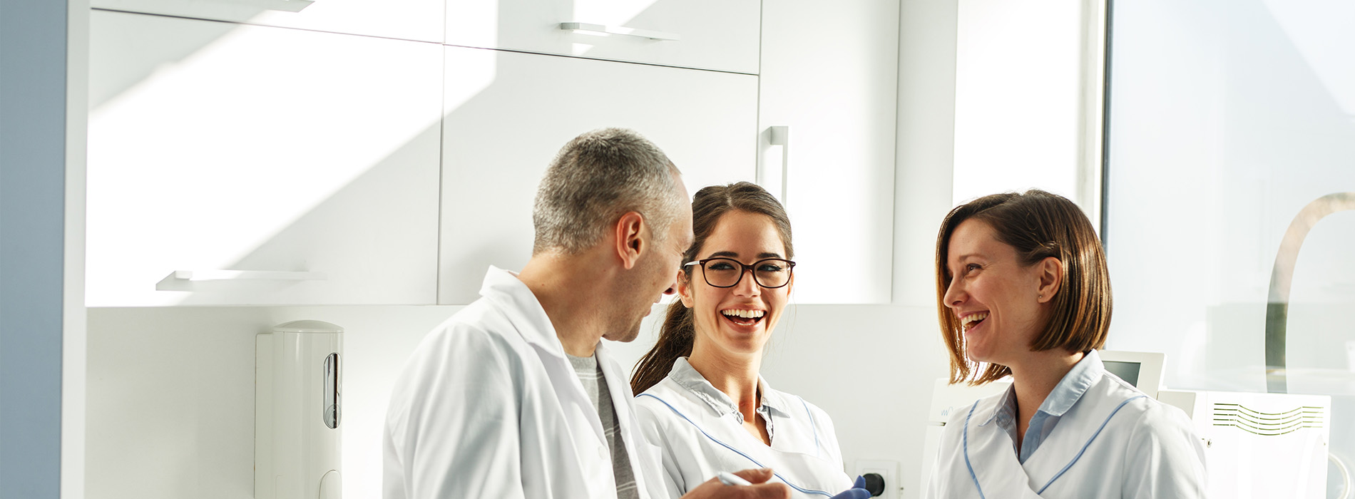 The image shows three individuals in lab coats standing together in a room with white walls, smiling and engaging in conversation.