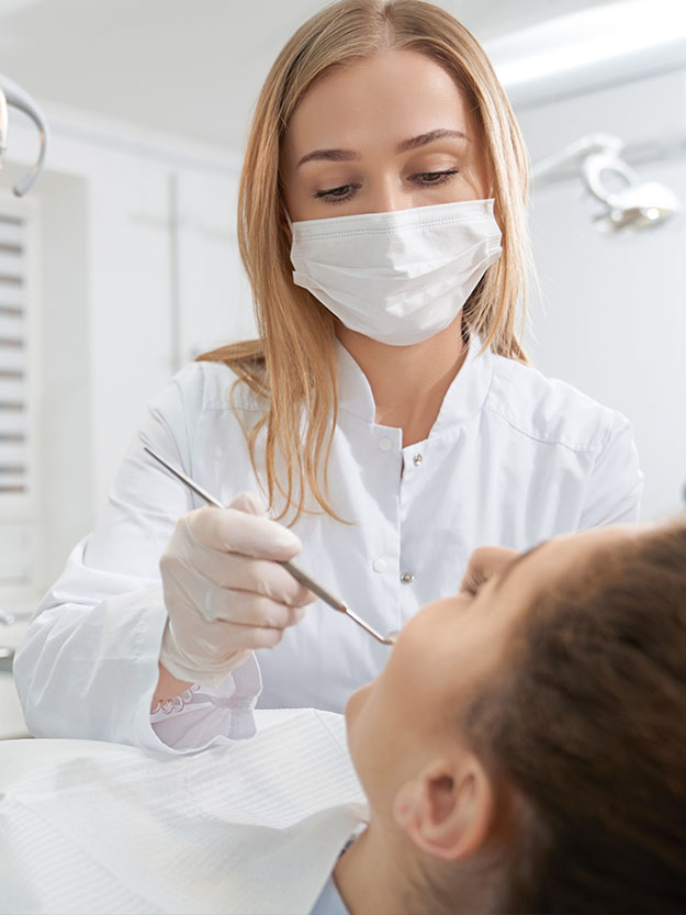 A dental hygienist wearing gloves and a mask is seated at a dental chair, performing work on a patient s teeth.