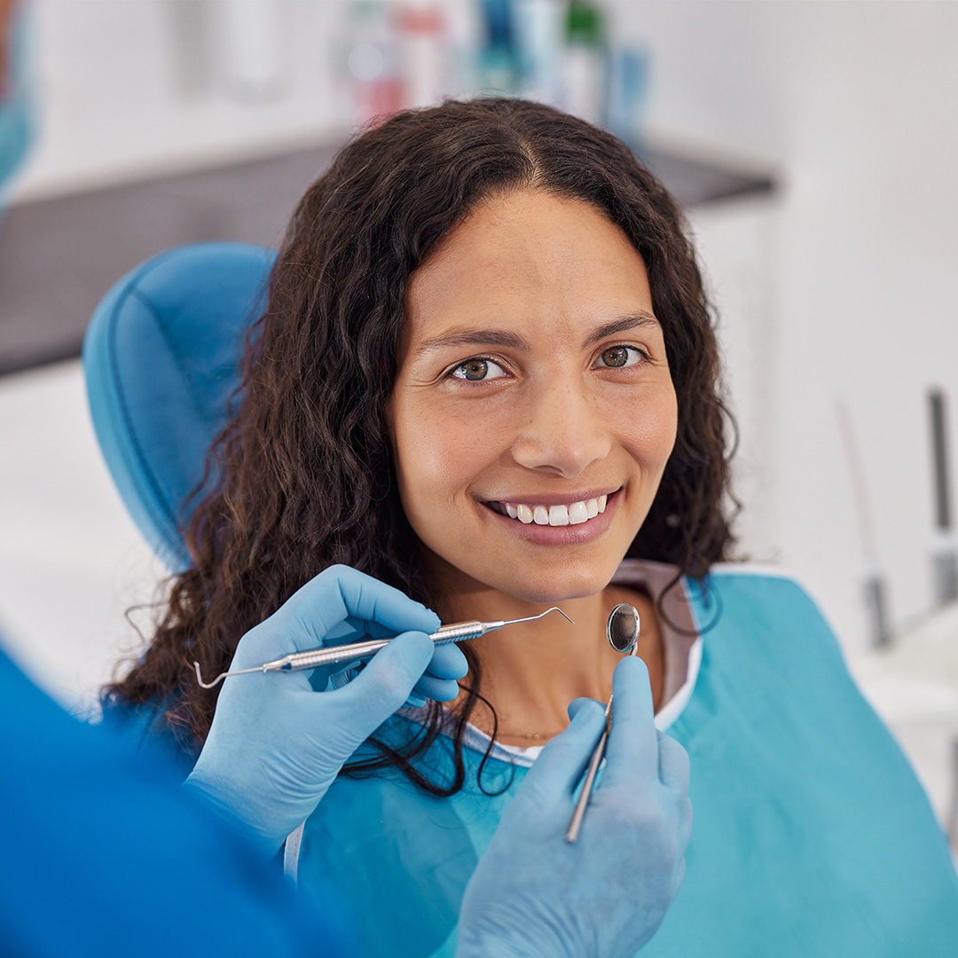 A woman seated in a dental chair, smiling at the camera, receiving dental care with a hygienist attending to her teeth.
