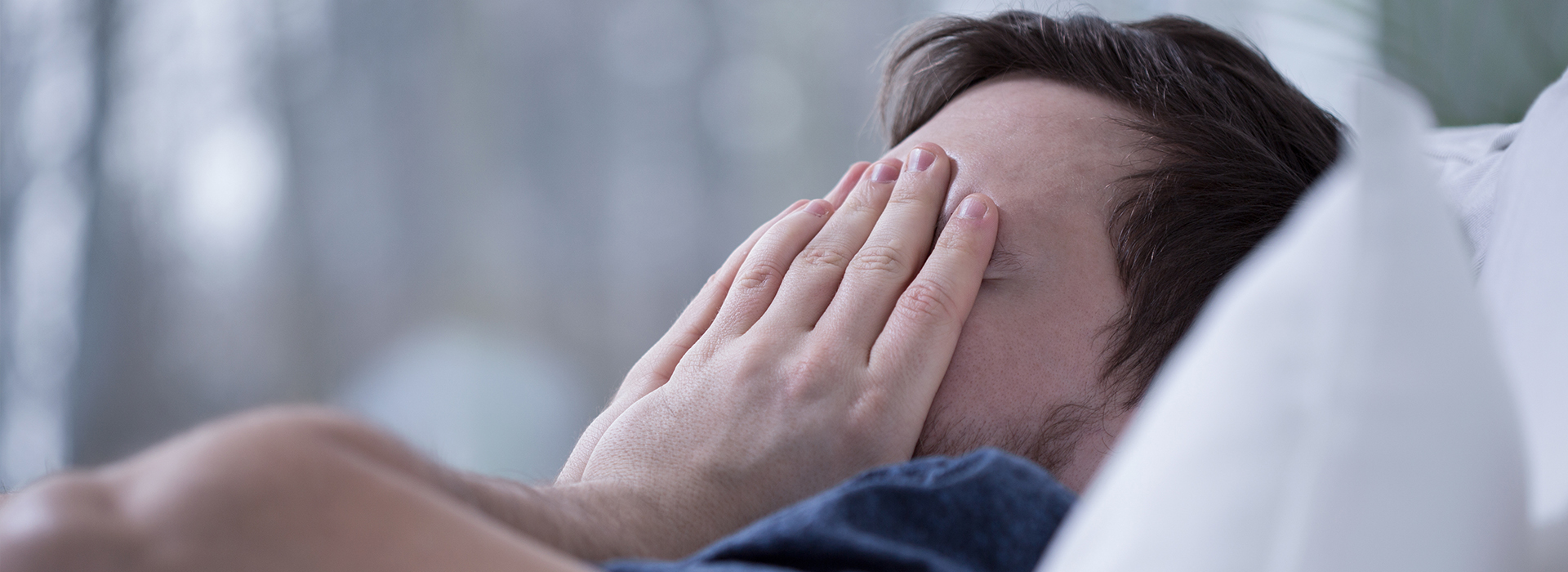 The image features a man lying on a bed with his head covered by his hand, possibly indicating discomfort or embarrassment.