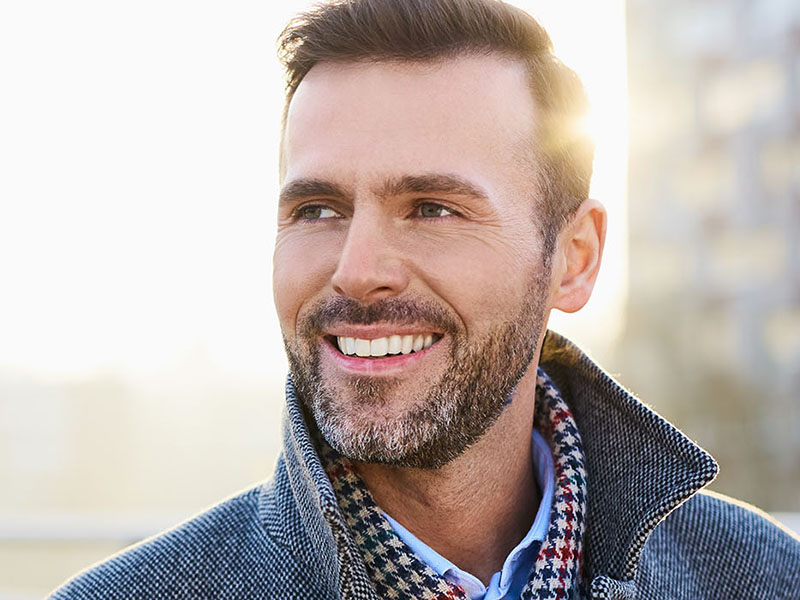 The image features a man with a beard, smiling at the camera, wearing a jacket and a checkered shirt, set against a blurred background with natural light.