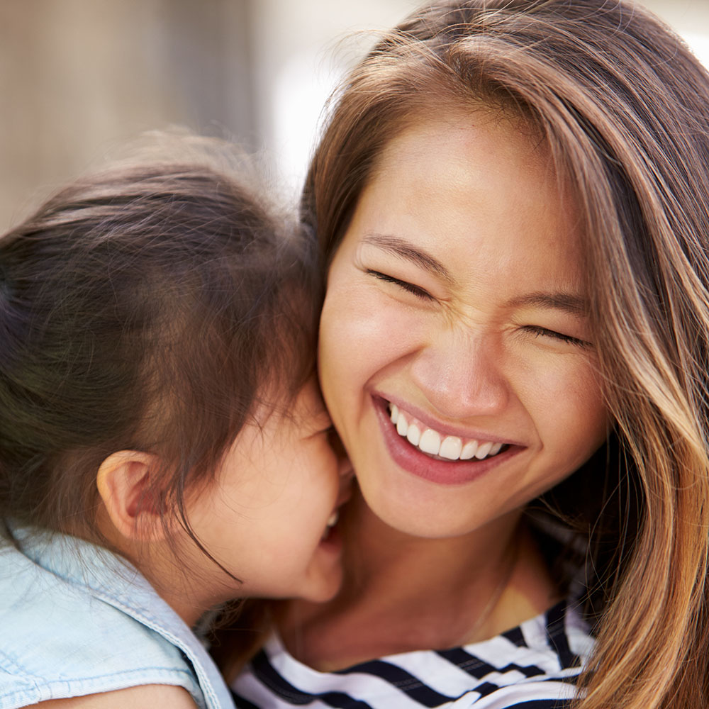 The image shows a woman with a young child, both smiling, sharing a tender moment in an outdoor setting during daylight hours.